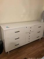 Front angled view showing the full white dresser with nine drawers, black handles, and visible scratches.