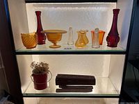Full view of 9 glass items arranged on a shelf under light including 5 flower vases in red and amber, 2 candy dishes, and 2 salt/pepper shakers.