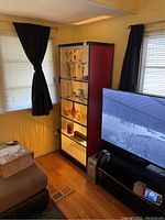 Full view of the lighted glass shelf unit in corner of room with black curtains and hardwood floor, showing the unit height relative to nearby TV and furniture.