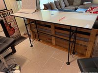Full view of the tall table showing the white top with plexiglass cover and black metal pipe frame standing on tiled floor next to seating area.