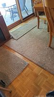Photo showing one large tan area rug on wooden floor beneath dining table and sliding glass door, with two matching tan door mats positioned nearby on floor.