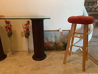 Wide view showing wooden stool with red cushion, two clear glass vases with red faux flowers on floor, and floral framed artwork behind panel leaning against wall.