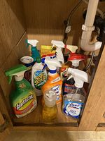 First photo showing mostly spray bottle cleaners including Fantastik, Soft Scrub, Lysol, Windex and other brands on a wood shelf under the sink