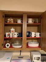 Cabinet with various coffee mugs, a decorative teapot, white ceramic pitcher, clear glass cups, and Pyrex baking dishes