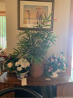 Wide shot showing six faux plants in various planters including straw, ceramic, and glass, arranged on a wooden dresser beneath framed artwork.