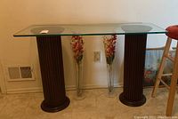 Front view of glass top console table showing clear glass panel on two dark brown fluted pedestal bases with round platforms. Floor vent and floral vases in background.