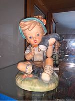 Front view of a Goebel porcelain figurine of a boy sitting with a blue hat, wearing traditional clothes including lederhosen and socks with polka dots, holding a pipe.