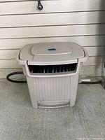 Close-up of beige Sun Cast hose caddy with water hose partially visible inside, placed on concrete floor against house siding.