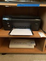 Black HP Photosmart multifunction printer placed on a wooden shelf with a sheet of paper partially inserted in the output tray.
