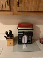 Kenmore Elity cordless electric kettle in box next to a wooden block holding six black-handled knives, small red and gray pouch on top of the box, and glass cutting block underneath with candle on right side.