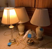 Four lamps arranged on carpet: one crystal base lamp with beige shade, two white ceramic base lamps with pleated shades, one small brass base lamp with glass prism shade
