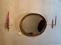 Photo of the oval wall mirror with two brass wall-mounted candle holders, each holding a red candle. Mirror shows ornate decorative bronze-toned frame.