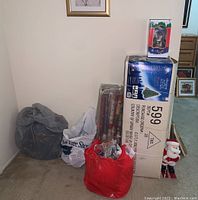 Photo of boxed Oregon pine tree and plastic bags containing Christmas decorations and wrapping paper rolls in corner of room.