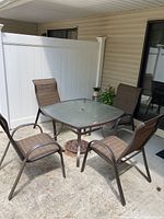 Patio set with square glass top table and four matching chairs arranged on concrete patio near white fence and siding wall.