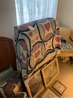 Folded single rollaway bed frame covered with patterned quilt, leaning against a wall with framed pictures around base.