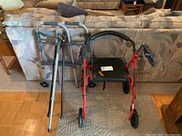 Three mobility aids shown together: a silver folding walker with wheels, a red rollator walker with seat and hand brakes, and a pair of silver crutches leaning against the couch.