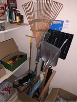 Photo showing various yard tools including metal rakes with wooden handles, black shovel, and large metal shovel.