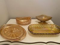 Four carnival glass items arranged on a table: one oval tray with handles and leaf petal pattern, one rectangular bowl, one square pedestal dish, one oval diamond pattern dish.