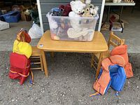 Overall view of children's wooden table, two chairs, life jackets on the backs, and plastic bin of stuffed animals on table.