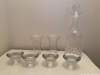 Photo showing one clear glass decanter with diamond-cut pattern and round stopper, two glass vases with floral embossed rims, and four small round candle holders with thick bases.