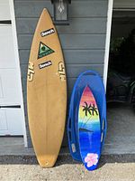 Photo showing a tan surfboard with Bench and Newsboys stickers standing next to a blue snow saucer decorated with tropical sunset, palm trees, and flower art.