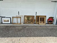 Lot of vintage pictures, frames and an ornate mirror leaning against a garage door outdoors.