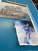 Two vintage vinyl posters, one showing a large sailing ship and the other a hockey player in blue and orange uniform on ice, both partially unrolled with some curling and wrinkles.