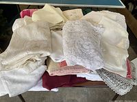 Stack of vintage white and cream textiles including lace tablecloths, towels, and bed sheets with various textures and patterns.