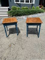 Pair of wooden side tables with natural wood tops and blue legs shown outdoors on pavement.