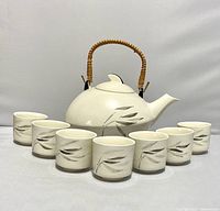Full set presentation of teapot centered with seven matching cups arranged in a row in front, showcasing hand-painted bamboo leaf design in gray on white ceramic.