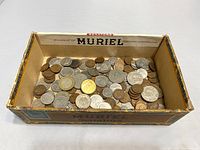Top-down view of the vintage Muriel cigar box bottom filled with assorted Canadian and Mexican coins of different metals and sizes.