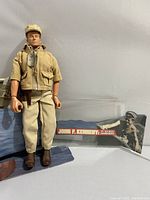 Front view of the John F. Kennedy G.I. Joe figure wearing tan jacket, dog tags, beige pants and brown boots. Display card with 'John F. Kennedy PT 109 Boat Commander' text and image included in background.