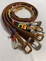 Seven belts coiled, showing variety of buckle styles and materials including leather and woven fabric.