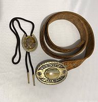 Top view of the tooled leather belt coiled next to the bolo tie with visible sandstone buckles.
