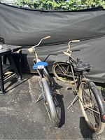 Two vintage bicycles outside on pavement against a black tarp, one silver and one black in color, showing rust and wear.