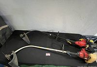 Overview of two gas-powered weed whackers side by side on black surface showing curved shafts, red motors, and yellow fuel tanks.