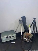 Photo showing the Argus automatic slide projector alongside two camera tripods in cases, the camera and its carrying case.