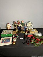 View of entire lot showing Byers Choice Caroler dolls, Santa figurines, wreath, ornaments, boxed decoration plaques, and assorted holiday decorations laid out on a black surface.