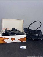 Photo of a white box with brown accents containing black leather boots and a black leather handbag placed beside the box on a black surface.