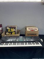 Photo showing electronic keyboard with two boxes of vinyl records behind it. Keyboard has multiple buttons, digital panel, and circular speakers on each side.
