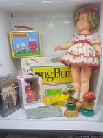 Overview of lot showing Shirley Temple doll, Peanuts lunchbox, Long Burn game, wooden toys, boxed figure, and VHS tape