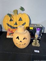 Photo showing ceramic pumpkin lantern with removable lid, large wooden jack-o'-lantern decoration, string pumpkin lights, Totally Ghoul inflatable box, and purple and gold skeleton candle holder on a black surface.
