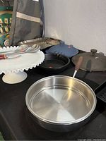 Wide view of kitchen supplies on a table showing a stainless steel mixing bowl, cookware lids, and frying pans.