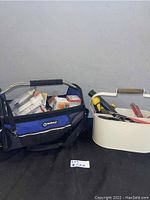 Photo showing Kobalt branded tool bag with metal handle and various boxed light bulbs visible inside, next to white toolbox containing hand tools.