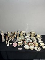 Overview of the collection showing multiple decorative statues, tea cups, plates, and small kitchenware laid out on a black tablecloth.