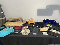 Overview of purses arranged on a black tablecloth; various colors and styles, some showing dirt and wear, some compact wallets or small bags, and also boxed umbrellas at the back of the table.