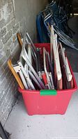 Red plastic bin filled with multiple folded metal frame folding chairs in a storage area with concrete floor and wall.