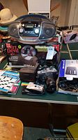 Photo showing the Sony radio, RCA CD player, Minolta and Studio 35 35mm cameras, Polaroid camera, Dirt Devil vacuum box, crossword puzzle books, and assorted cables.