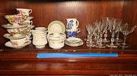 Stacked teacups and saucers with various floral patterns, small bowls and plates behind, alongside clear etched stemware glasses