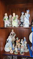 Photo of multiple vintage figurines arranged on wood shelves, including children and couples in period clothing, various sizes and colors